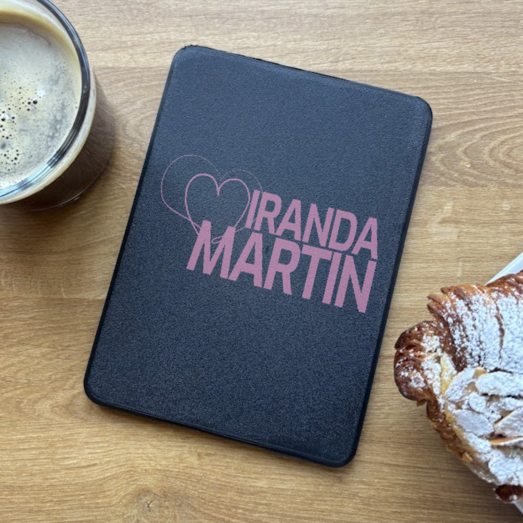 Black kindle case with 'MIRANDA MARTIN' logo on a wooden surface with a cup and pastry.