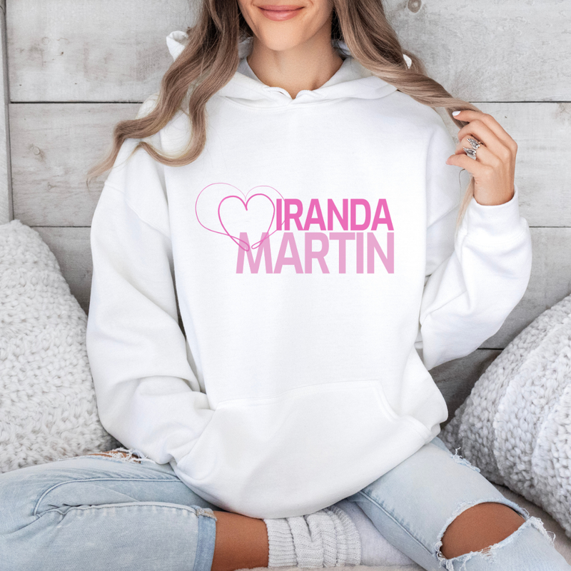 Person wearing a white hoodie with 'MIRANDA MARTIN' printed on it, sitting against a wooden wall.