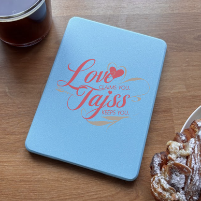 Light blue notebook with red cursive text 'Love Claims You. Tajiss Keeps You.' and a heart icon.