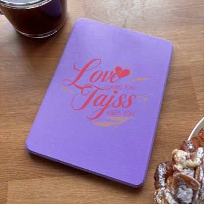 Purple notebook with red cursive text 'Love claims you, Tajss keeps you' on a wooden table.