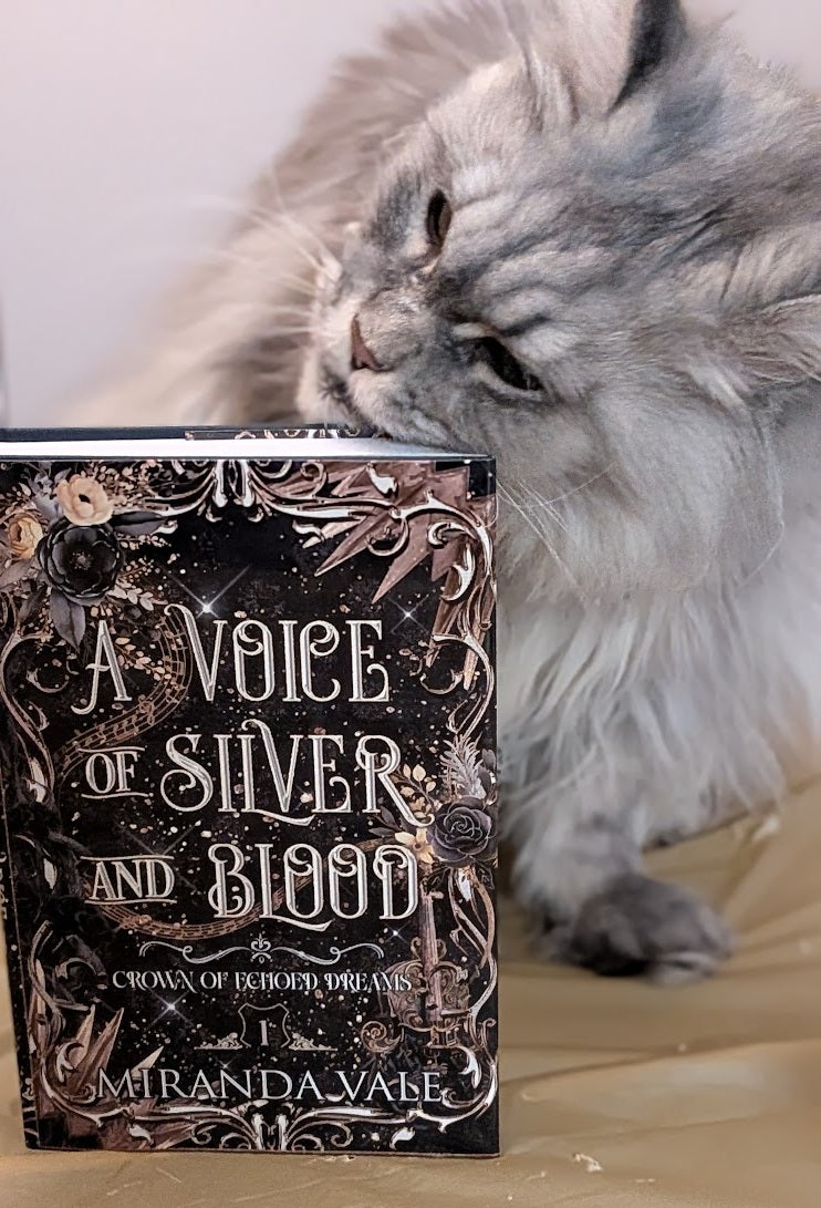 Cat sniffing a book titled 'A Voice of Silver and Blood' by Miranda Vane on a wooden surface.